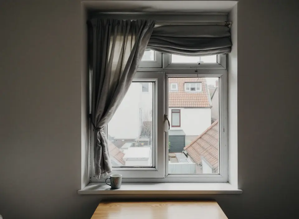 Een slaapkamerraam met witte kozijnen, een opzijgebonden gordijn en een mok in de vensterbank, met uitzicht op achtertuinen en pannendaken