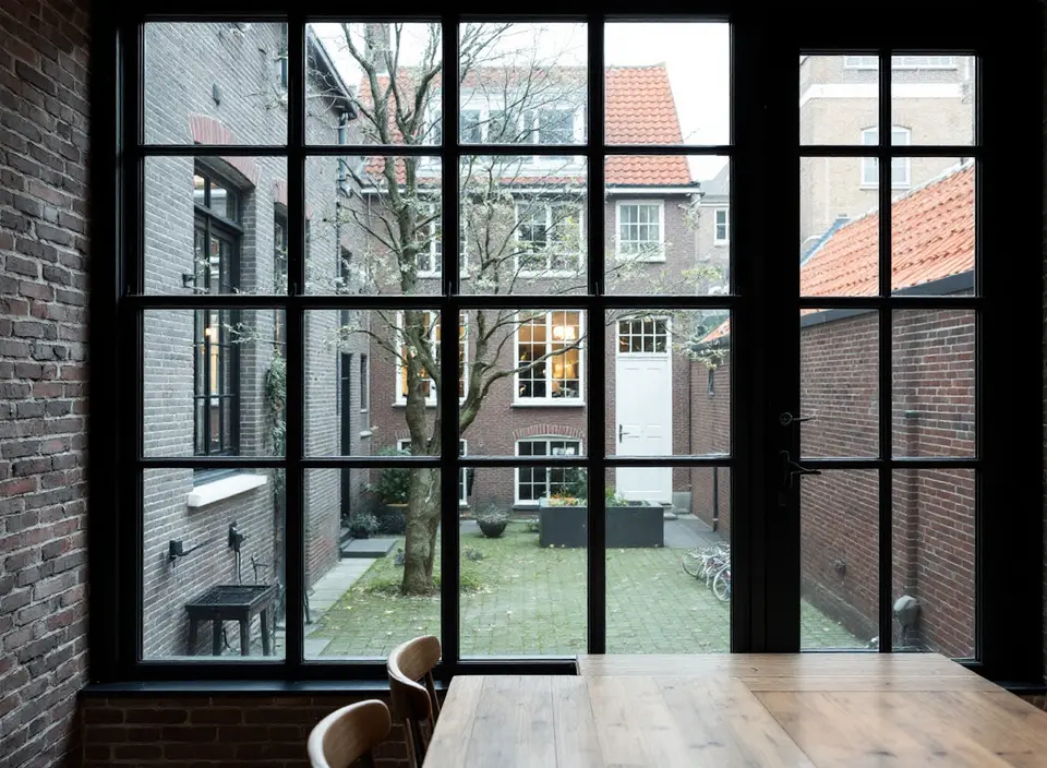 Een groot zwart stalen raam met kleine ruiten in een verbouwd pakhuis, met een houten tafel ervoor en uitzicht op een binnenplaats